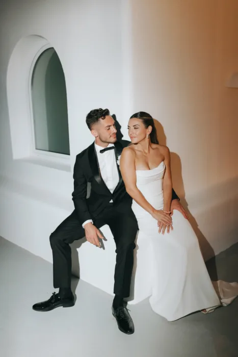 The couple sit beneath curved white architecture, lit by warm evening flash.