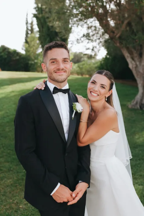 The couple smile together on the lawn, framed by soft greenery and afternoon light.