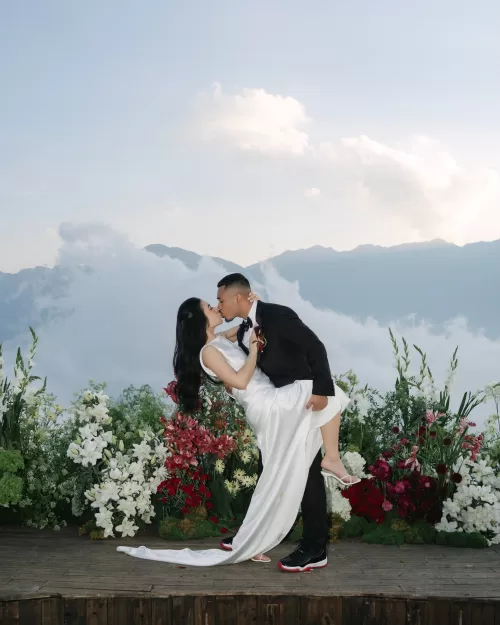 The couple kissing on a floral stage with clouds and blue mountain silhouettes behind them.