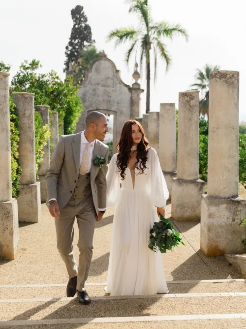Bride and groom walking between historic stone columns at Pousada Palácio Estoi