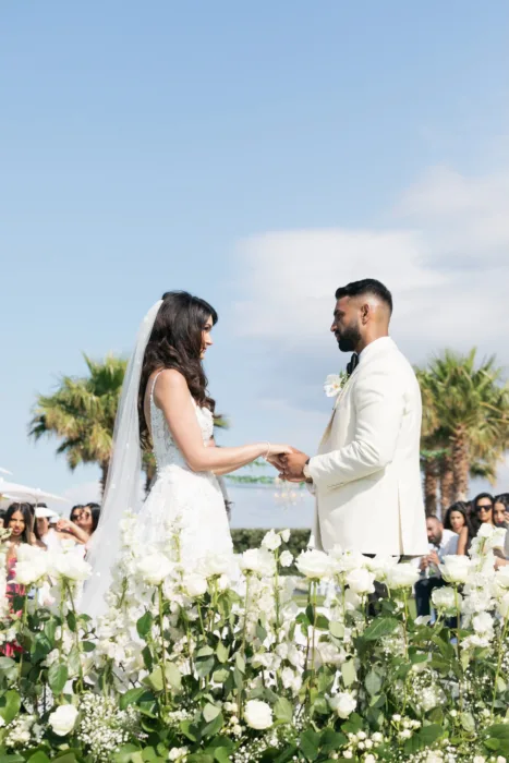 Couple stands among white roses and palms during their outdoor ceremony.