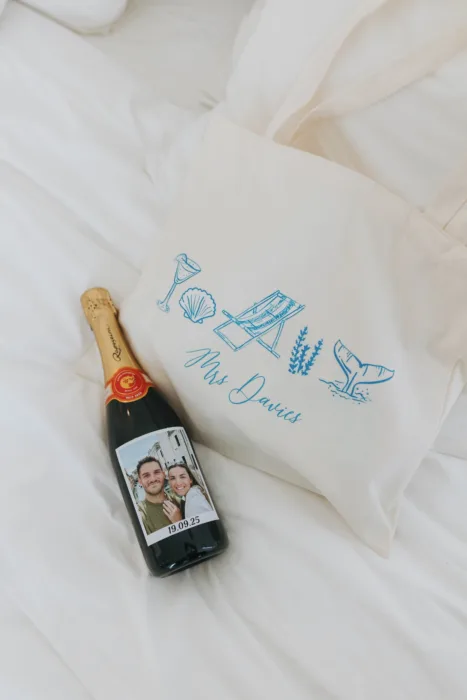 A personalised tote bag and bottle of cava rest on crisp white bedding.