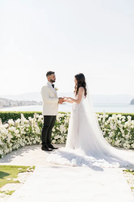 Bride and groom exchange vows before white flowers and an open sea backdrop.