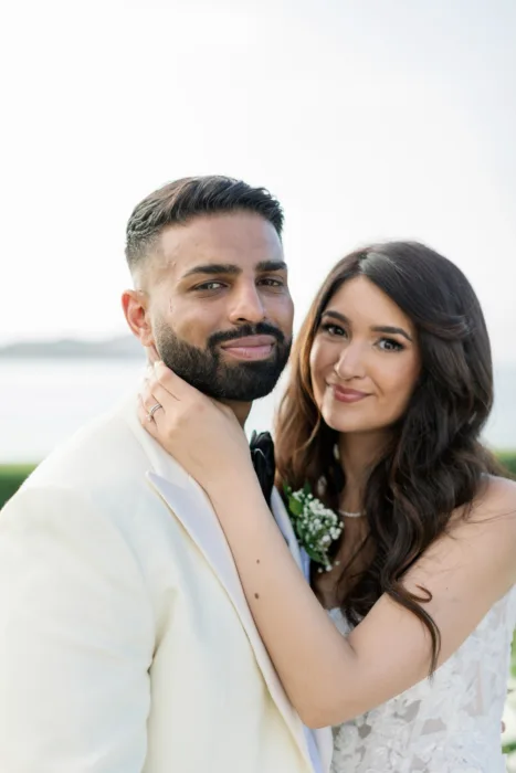 Newlyweds pose closely together with the soft blue sea behind them.