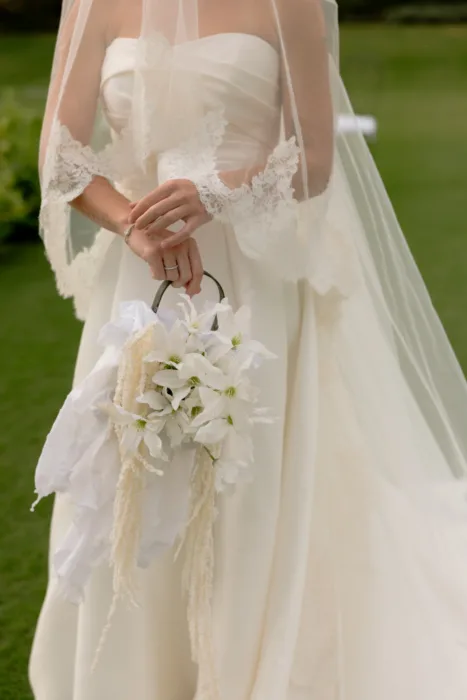 Bride holding a handled bouquet of white blooms with trailing cream amaranthus against her satin gown