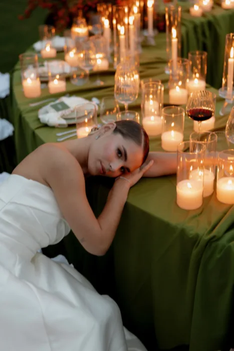 Bride resting her head on her arm among pillar candles and wine glasses on a green table
