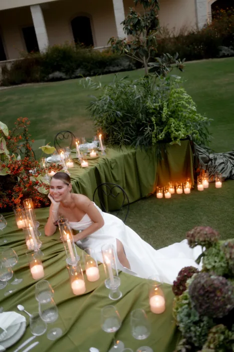 Smiling bride resting her chin on her hand at a candlelit olive-green table, hydrangeas spilling beside her