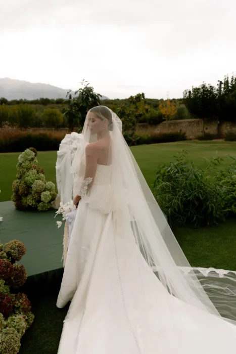 Bride in profile beneath a cathedral-length veil, the lace trailing across the lawn toward the hills