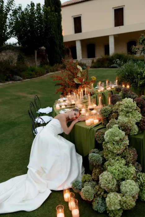 Bride leaning on the candlelit table at dusk, her full satin skirt pooling on the grass
