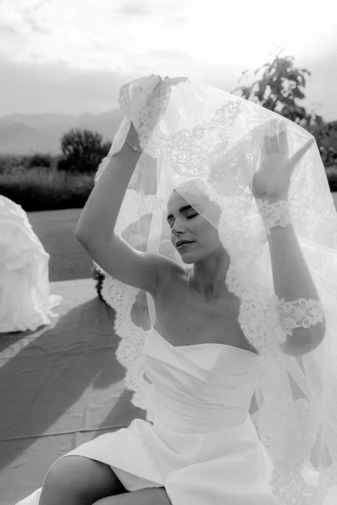 Black and white portrait of the bride lifting her lace-edged veil, eyes closed, mountains behind