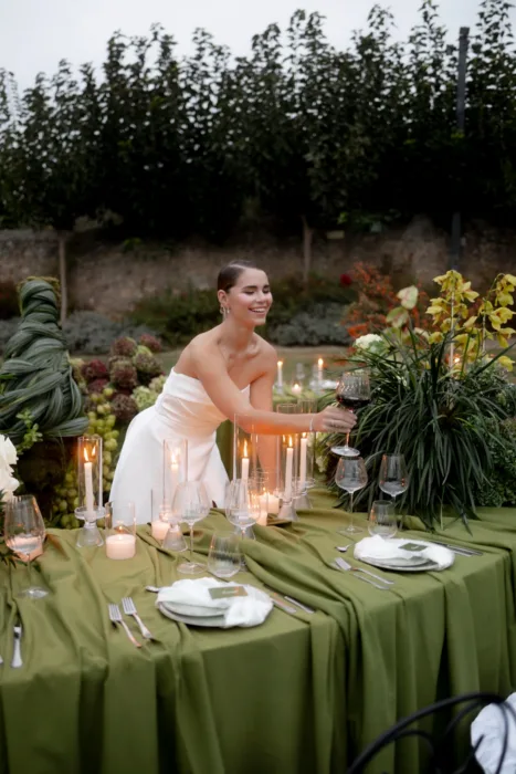 Laughing bride in a strapless gown reaching across the table with a glass of red wine