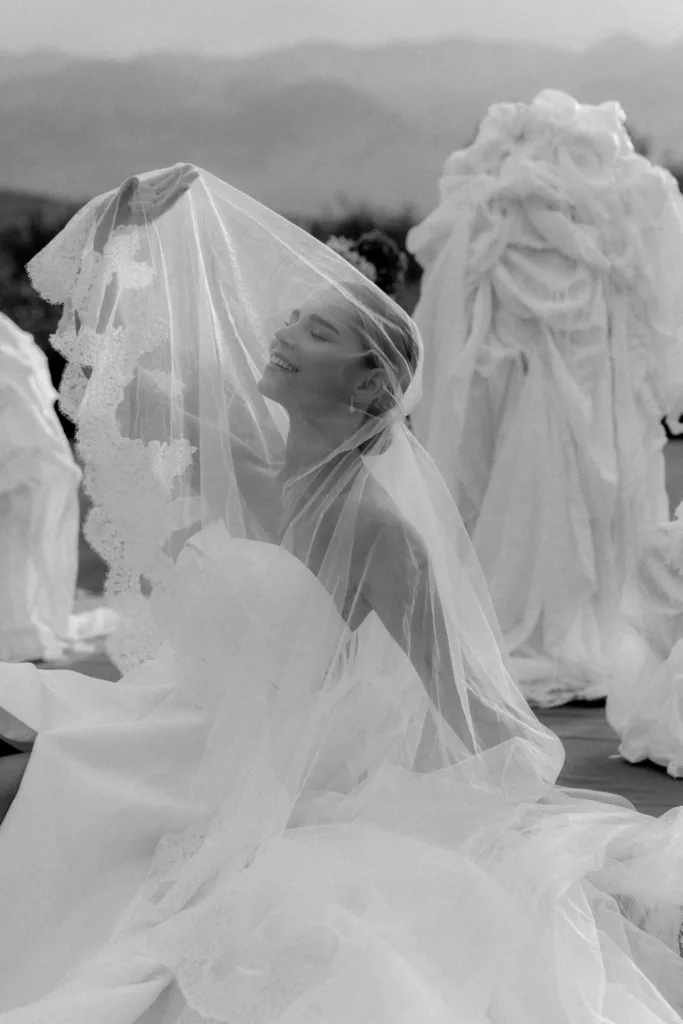 Laughing beneath a sheer veil, the bride sits among draped white sculptures in soft monochrome