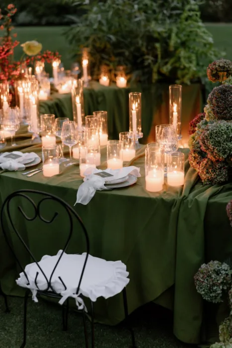 Hurricane candles and crystal glassware glowing along a moss-green table as evening falls