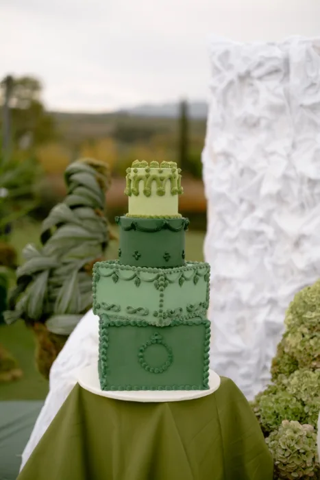 Close-up of a four-tier green cake with ornate piping, set on olive linen in the garden
