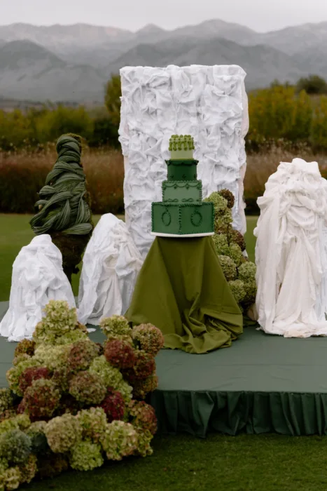 Vintage-style tiered cake in shades of green, framed by draped white sculptures and Cretan mountains