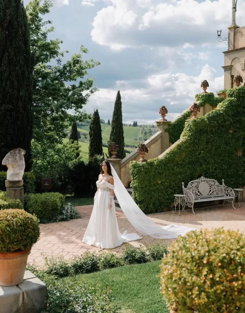 A bride in an elegant gown with a long veil walking through the ivy-covered courtyard of Tenuta Corbinaia, surrounded by Tuscan cypress trees.