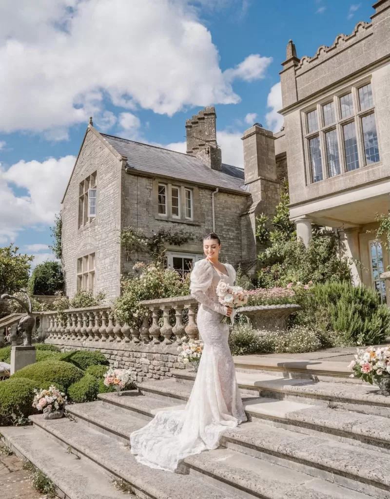 The bride poses on stone steps in front of the grand Euridge Manor, holding a blush-toned bouquet under sunny skies.