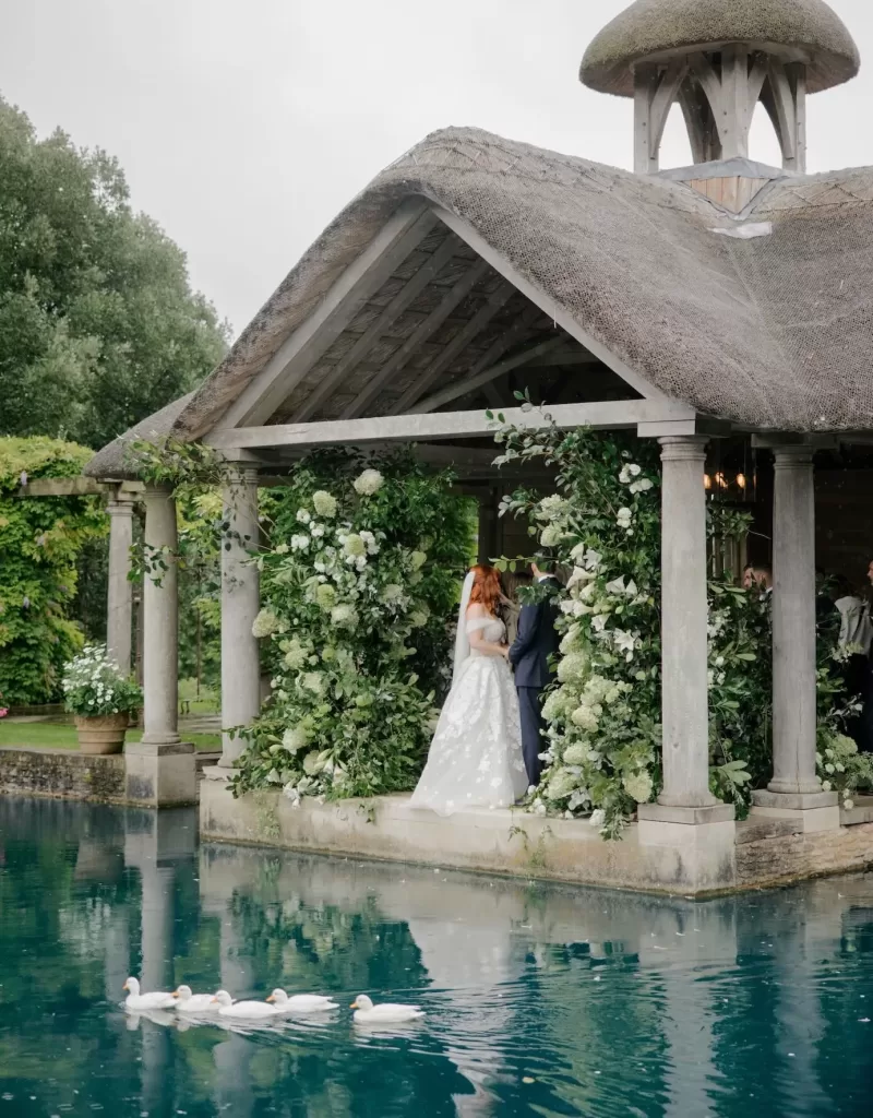 The couple exchanges vows beneath the pavilion at Euridge Manor, while a line of ducks glides across the water.