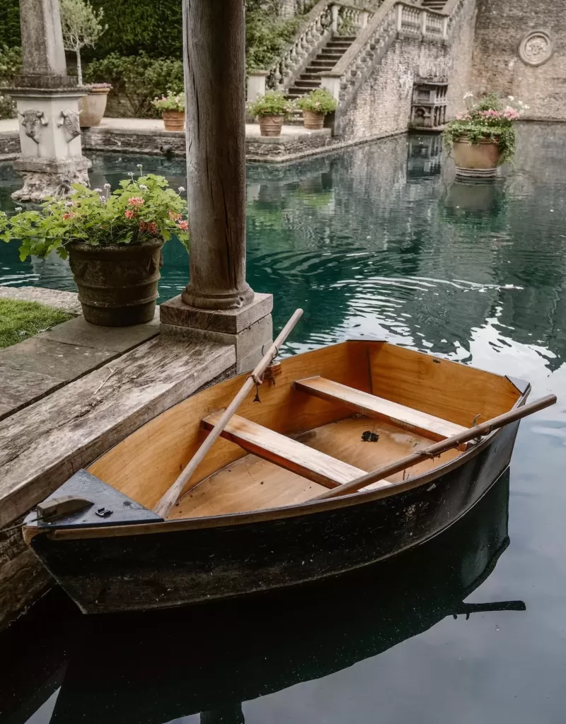 A wooden rowboat floats gently on the still turquoise pond at Euridge Manor, surrounded by stone columns and lush greenery.