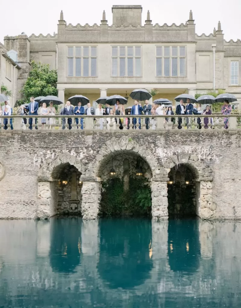 Wedding guests holding umbrellas on the stone terrace at Euridge Manor, overlooking the turquoise water below.