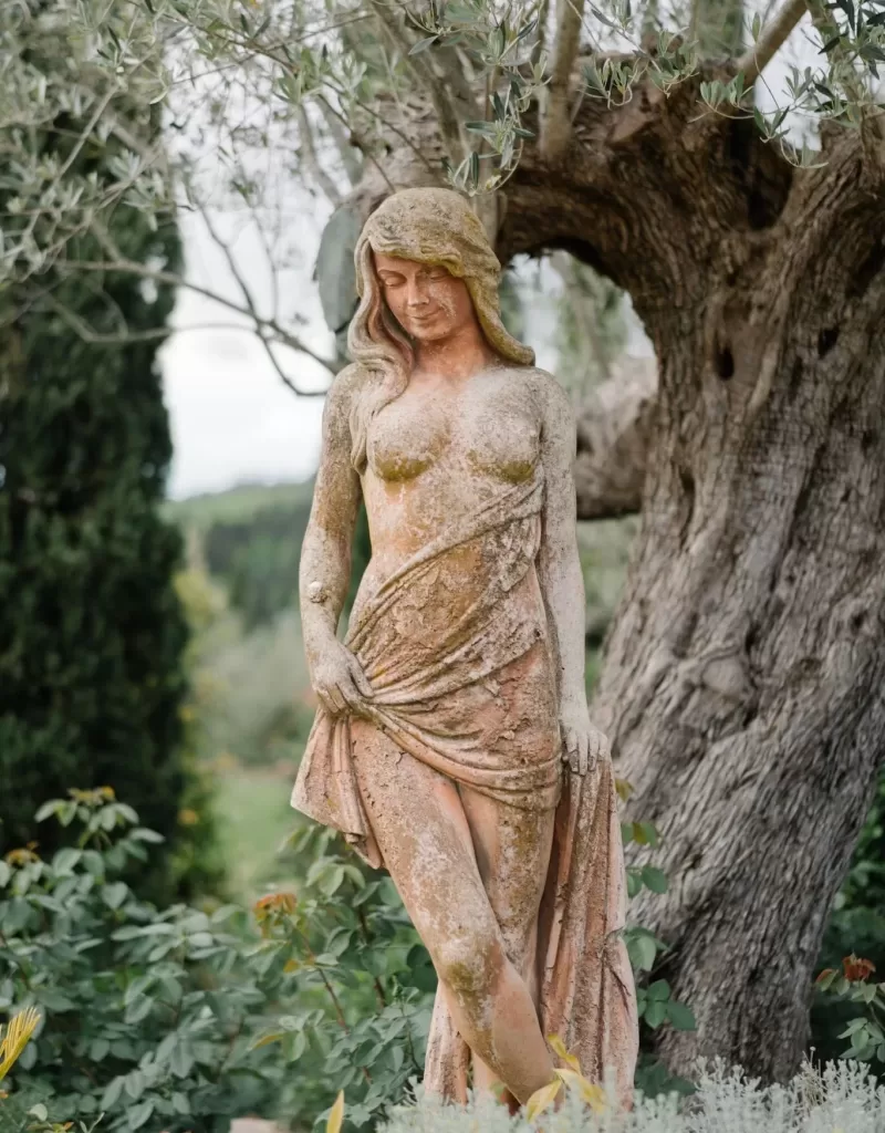 A weathered stone statue of a woman standing beneath an olive tree at Tenuta Corbinaia, adding charm to the Tuscan garden setting.
