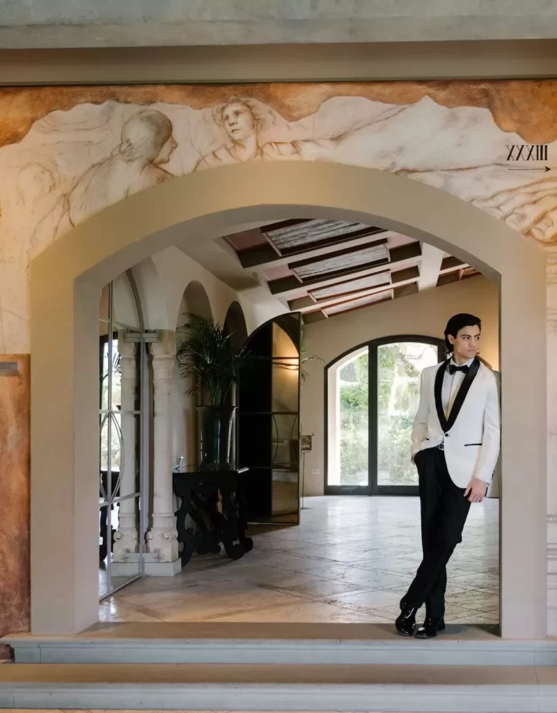 Groom in a white tuxedo standing in the frescoed archway of Tenuta Corbinaia, blending modern style with Italian heritage.
