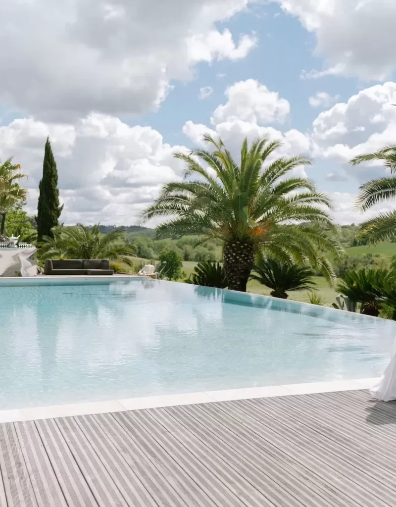 Bride in a white robe walking beside the palm-fringed pool at Tenuta Corbinaia with rolling Tuscan hills in the distance.