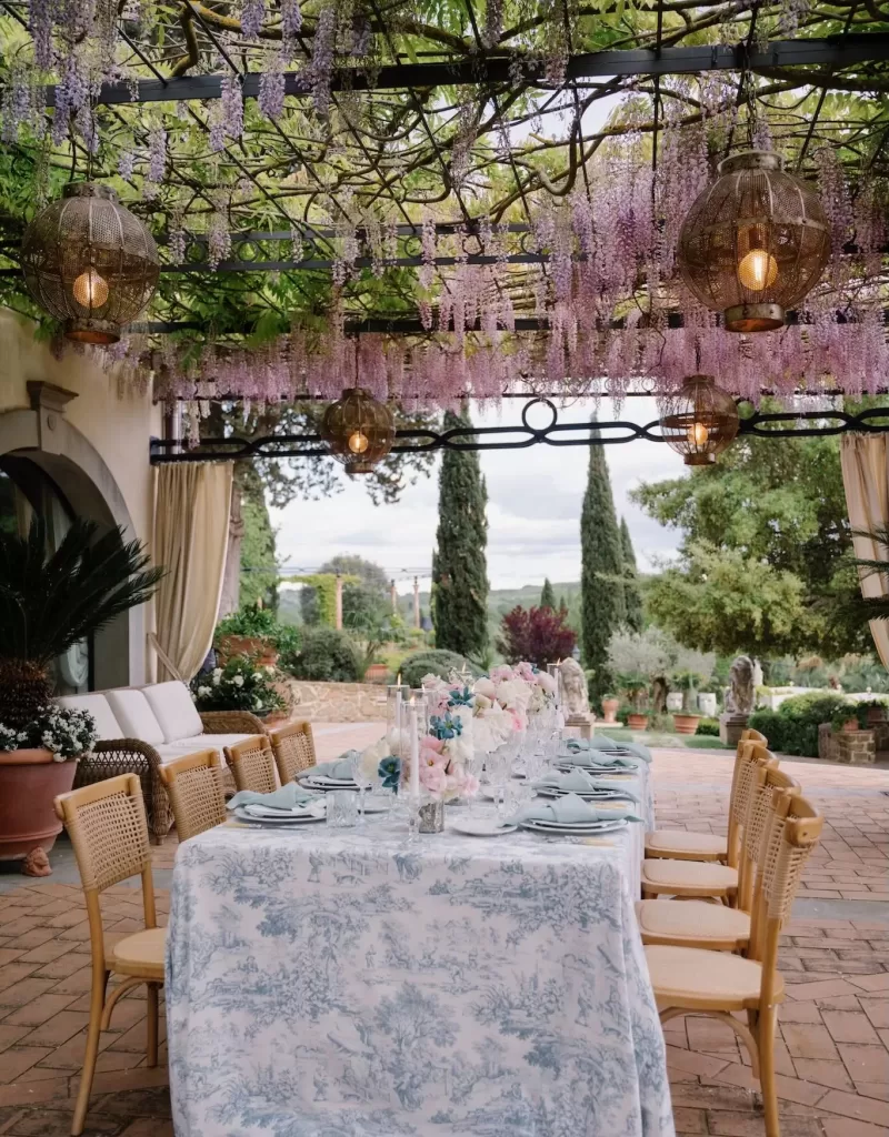Long reception table under blooming wisteria pergola at Tenuta Corbinaia with views of Tuscan gardens.