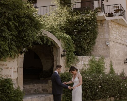 Bride and groom under the stone archway of Vasilias Nikoklis Inn, framed by olive trees and warm Mediterranean light.