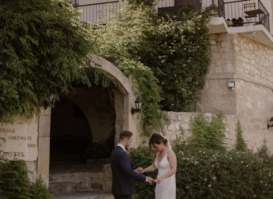 Bride and groom under the stone archway of Vasilias Nikoklis Inn, framed by olive trees and warm Mediterranean light.